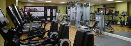 Cardio and weight machines line up, standing idle inside a well-lit gym; treadmills, ellipticals, cable station, benches, and bikes face mirrors and televisions on polished wood floors.