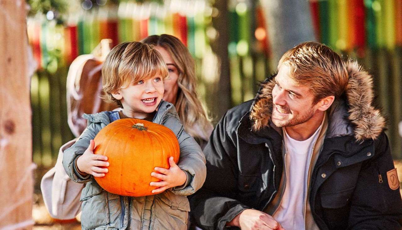 Child holds a bright orange pumpkin and smiles, gripping it with both hands. Nearby adults watch. Outdoors in autumn, bundled in jackets, with blurred trees and colorful ribbons in background.