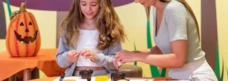 Two people craft decorations, folding small papers and using tools on an orange-covered table; in a festive room with jack-o’-lantern, green stalk shapes, and warm colors suggesting a Halloween-themed activity.
