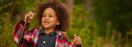 Child holds a stick, smiling and gesturing, in a forest clearing. Wearing a red plaid jacket over denim, they stand amid green foliage with soft, natural light and blurred background.