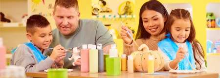 Children and adults paint small ceramic figures, dipping brushes into colorful paint bottles, seated at a table; bright craft studio context with shelves and decorations, pumpkin centerpiece, and aprons worn.