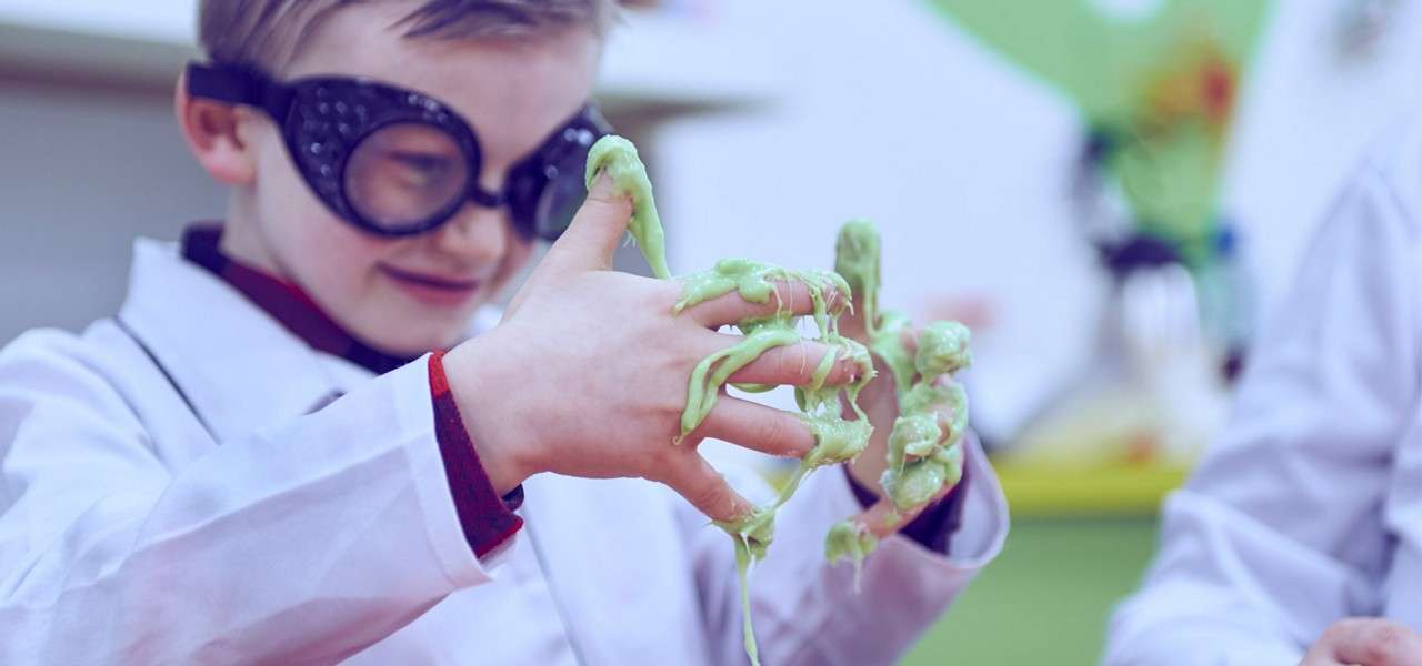 Child in oversized goggles manipulates gooey green slime, stretching it between sticky fingers, while wearing a white lab coat in a bright classroom or play-lab setting.