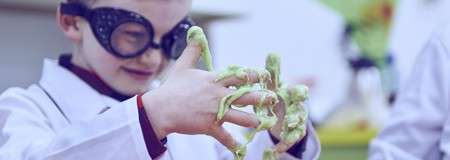 Child scientist squeezes gooey green slime between fingers, stretching strands. In a classroom or lab setting, wearing protective goggles and white lab coat, another child partially visible beside them.