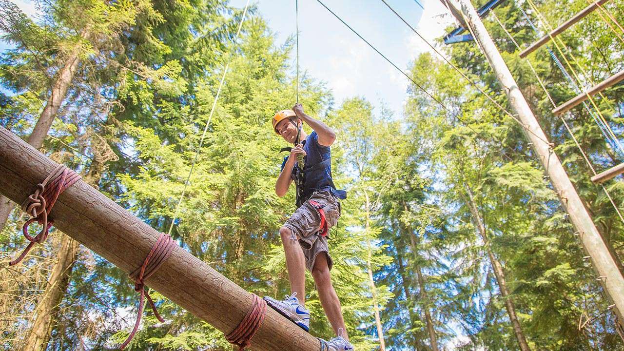 Person in helmet and harness balances on a suspended log, gripping a safety rope, within a high ropes course among tall evergreen trees and cables.