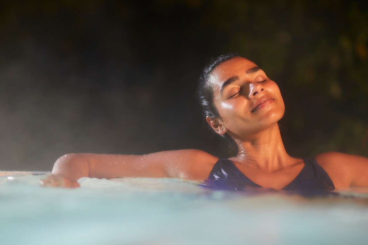 Person floating with arms outstretched, eyes closed, smiling softly; enjoying warm water. Soft steam rises around them in a dimly lit pool, suggesting nighttime relaxation outdoors.