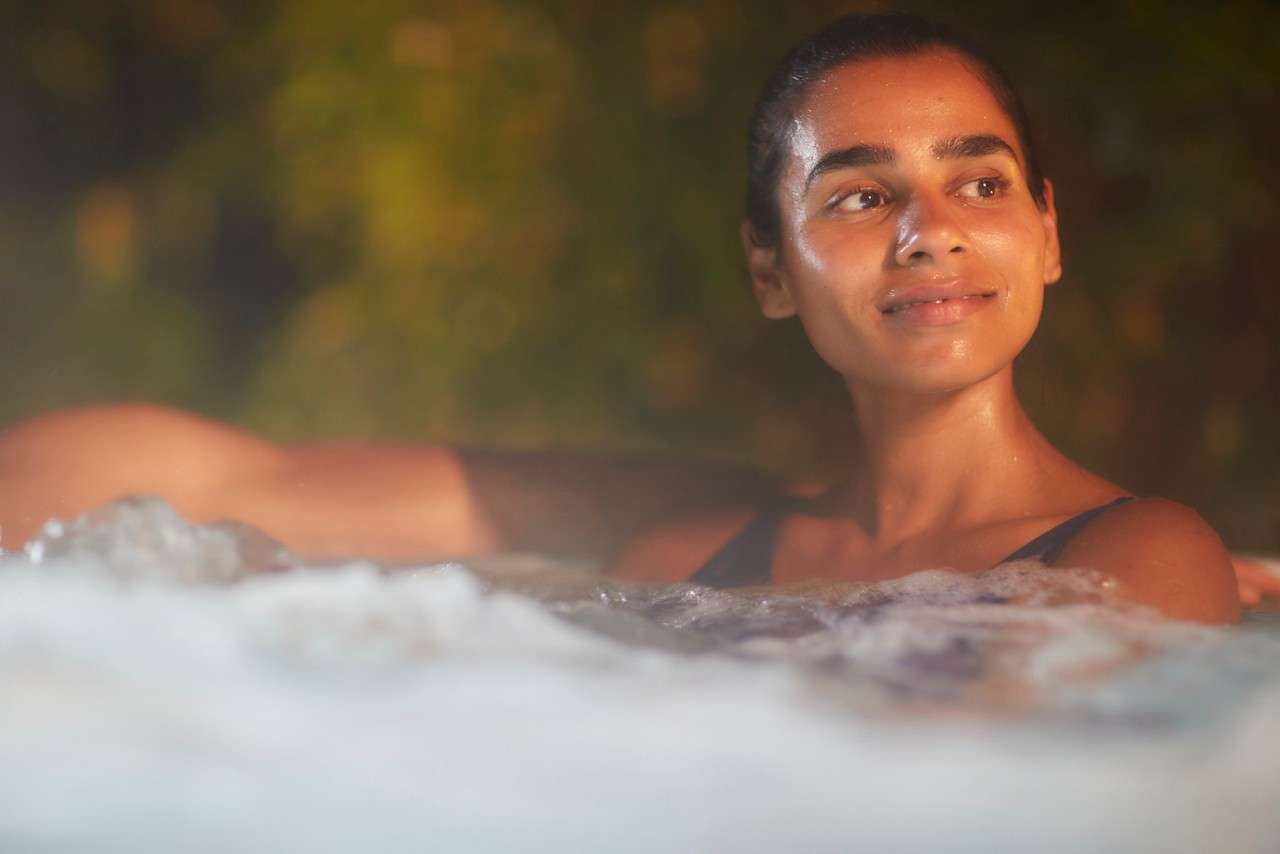 A smiling person relaxes in a bubbling hot tub at night, leaning back with an arm on the edge; warm light on their face; out-of-focus greenery in the background.