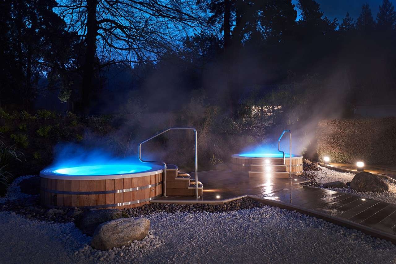 Two round hot tubs emit blue glow and steam at night, set on wooden decks amidst pebbled ground, rocks, handrails, and silhouetted trees in a landscaped garden.