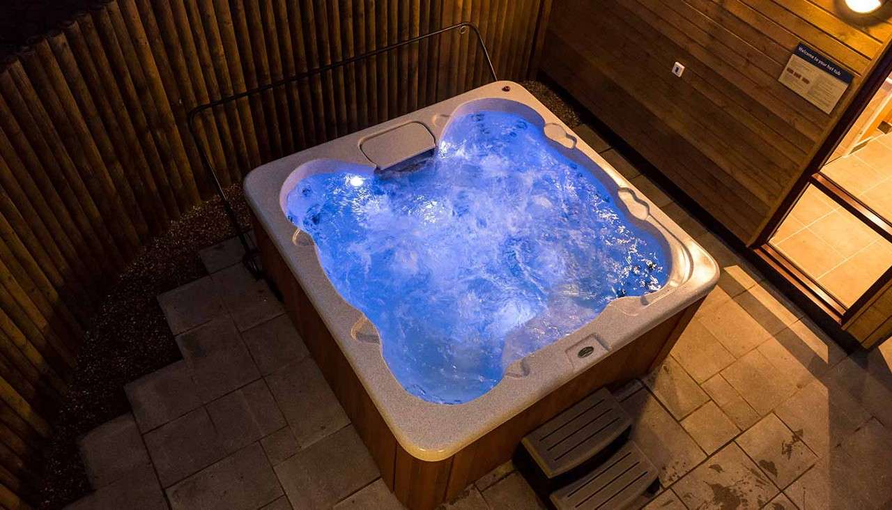 Hot tub bubbles vigorously, illuminated blue, with built-in jets churning. It sits on a tiled patio beside wooden walls and fencing, with a small step and handrail, viewed at night.