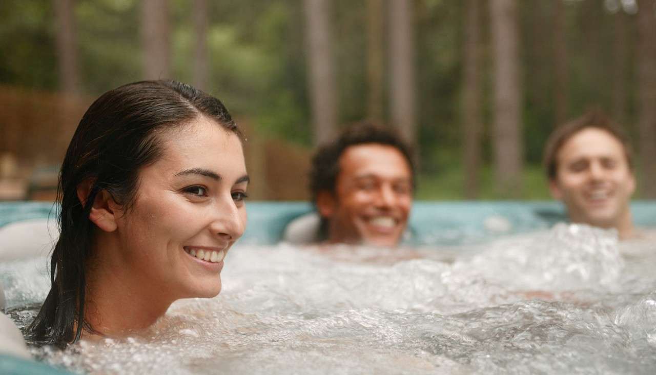 Three people relax, soaking in a bubbling hot tub, smiling and chatting, surrounded by an outdoor setting with trees in soft focus.