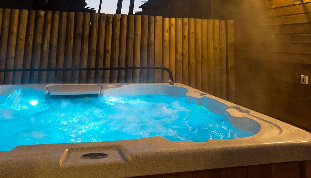 Hot tub bubbles vigorously, blue underwater lights glowing, steam rising. Set outdoors on a deck, bordered by vertical wooden fence and siding, with a grab bar and control panel visible.