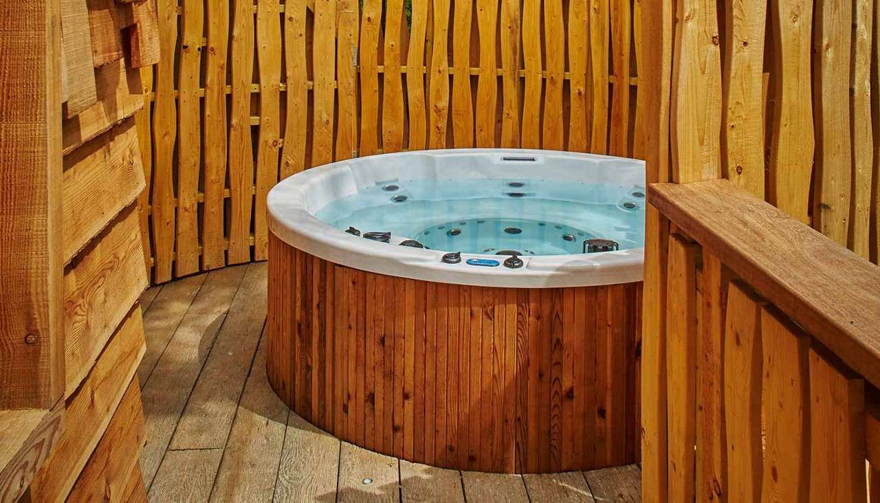 Round hot tub containing clear water sits idle, jets visible, within a small wooden enclosure and deck, surrounded by vertical cedar slats and a handrail.