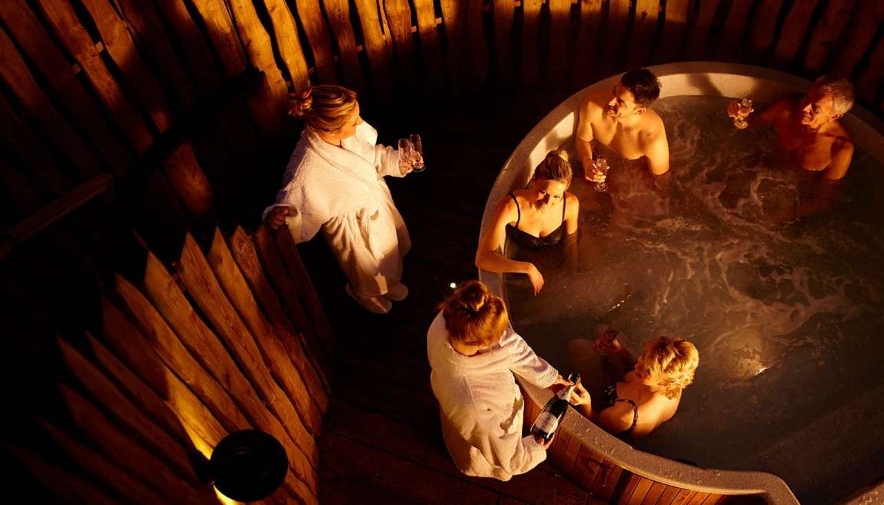 Group in a circular hot tub relaxes and toasts with drinks, while two people in robes pour wine; surrounded by wooden walls and warm lighting, creating a cozy spa setting.