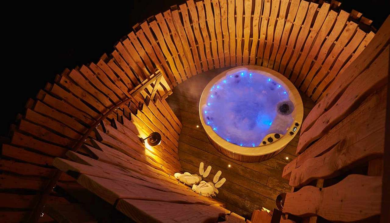 Hot tub bubbles gently, glowing with blue lights, inside a circular wooden enclosure at night; robes and slippers lie on the deck nearby, warm lamp illuminating rustic planks.
