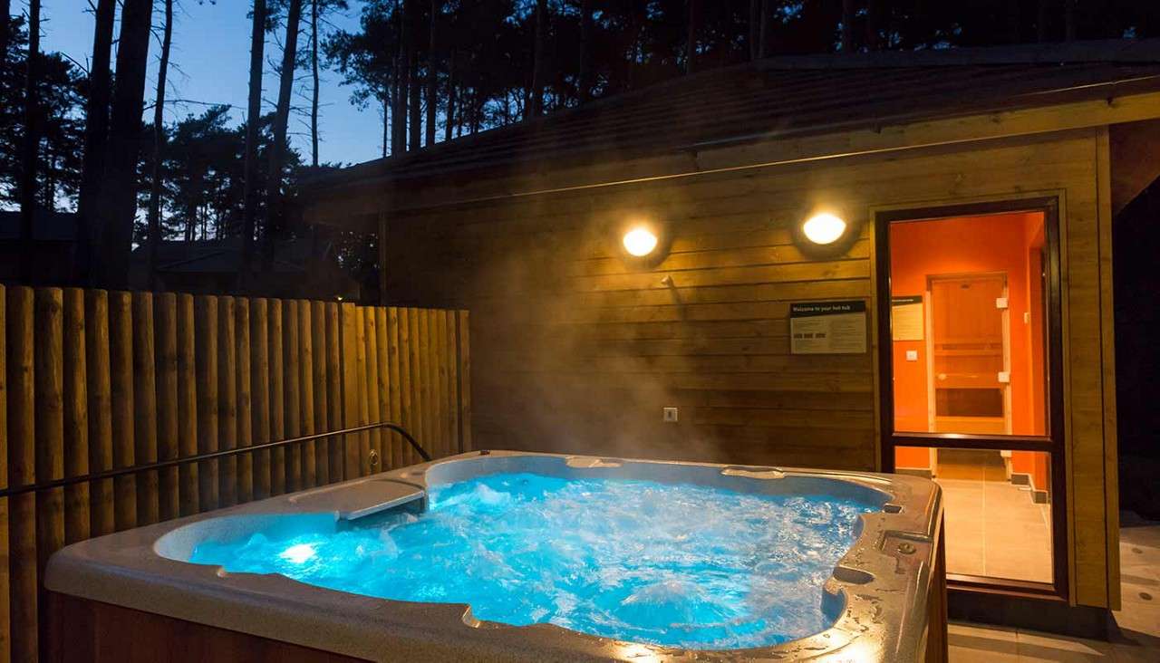 Hot tub bubbles and steams, glowing blue. It sits on a fenced outdoor deck beside a lit wooden cabin at dusk, with pine trees silhouetted in the background.