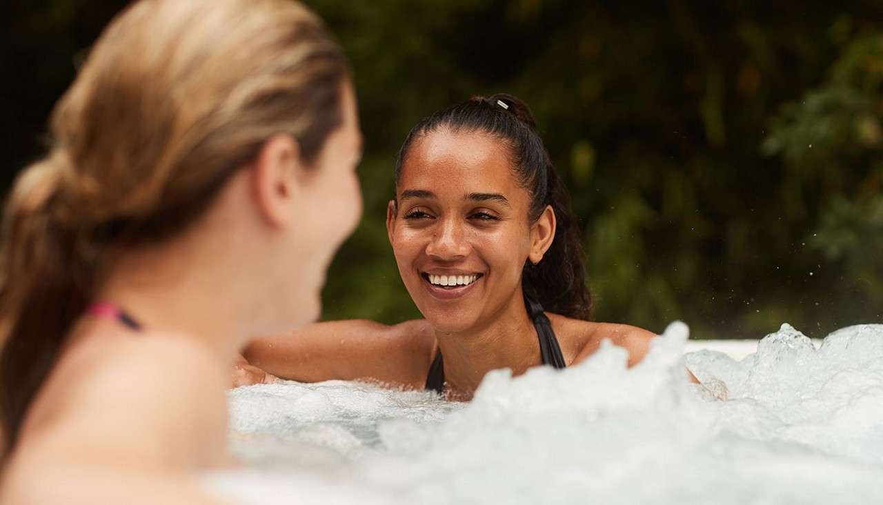 Two people smile and chat while soaking in a bubbling hot tub; the nearer person leans on the edge, slightly blurred, as the other faces them; outdoor setting with leafy greenery background.