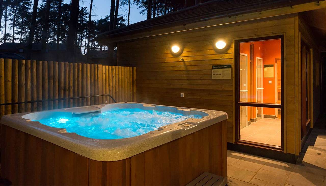 Hot tub bubbles vigorously, glowing blue and steaming, beside a wooden cabin with lit doorway and wall lights, enclosed by a fence beneath dusk sky and pine trees.