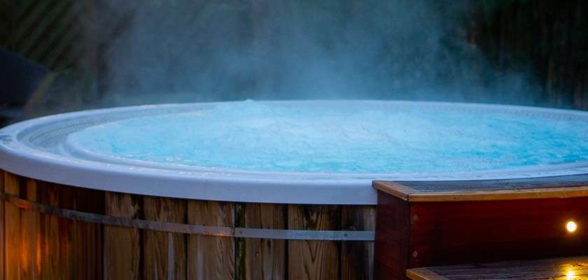 Round outdoor hot tub bubbles vigorously, steam rising. Context: wooden barrel-style sides, small lit steps nearby, dim evening yard with dark foliage.