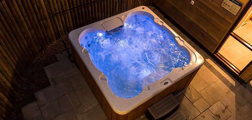Hot tub bubbles vigorously, glowing blue, within a square patio nook; wooden fence and wall surround it, steps sit beside the rim, and tiles and gravel form the outdoor setting.