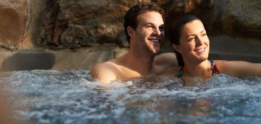Two adults relax, smiling, while soaking in a bubbling hot tub; water churns around them as they lean by a stone-lined pool wall, likely outdoors under warm light.