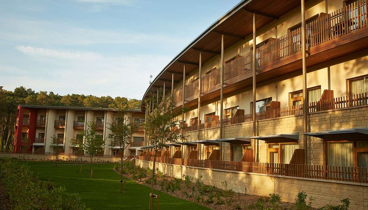 Curved multi-story hotel building displays rows of wooden balconies and pillars, overlooking a landscaped lawn with young trees; warm evening light and surrounding forest create a tranquil resort setting.