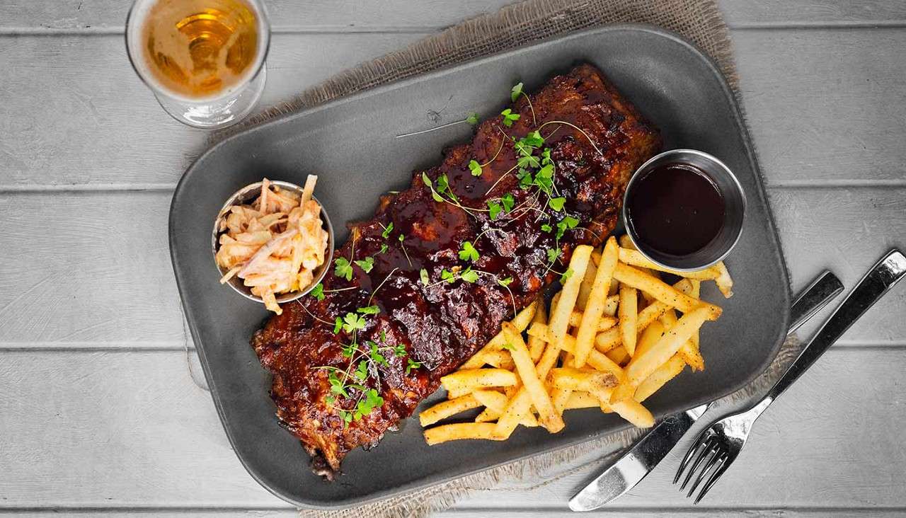 Rack of barbecue ribs sits on a platter, garnished with microgreens, with fries, coleslaw, and dipping sauce; nearby, a beer glass and cutlery rest on a wooden table.