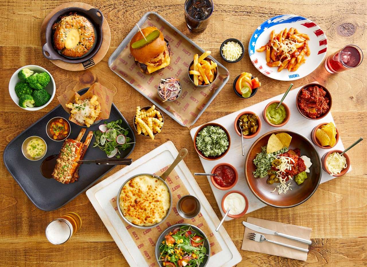 Assorted dishes—burger with fries, pasta, ribs, salads, dips, broccoli, and drinks—are arranged on a wooden table. Text: HUCKS (repeated on tray liners and one plate).