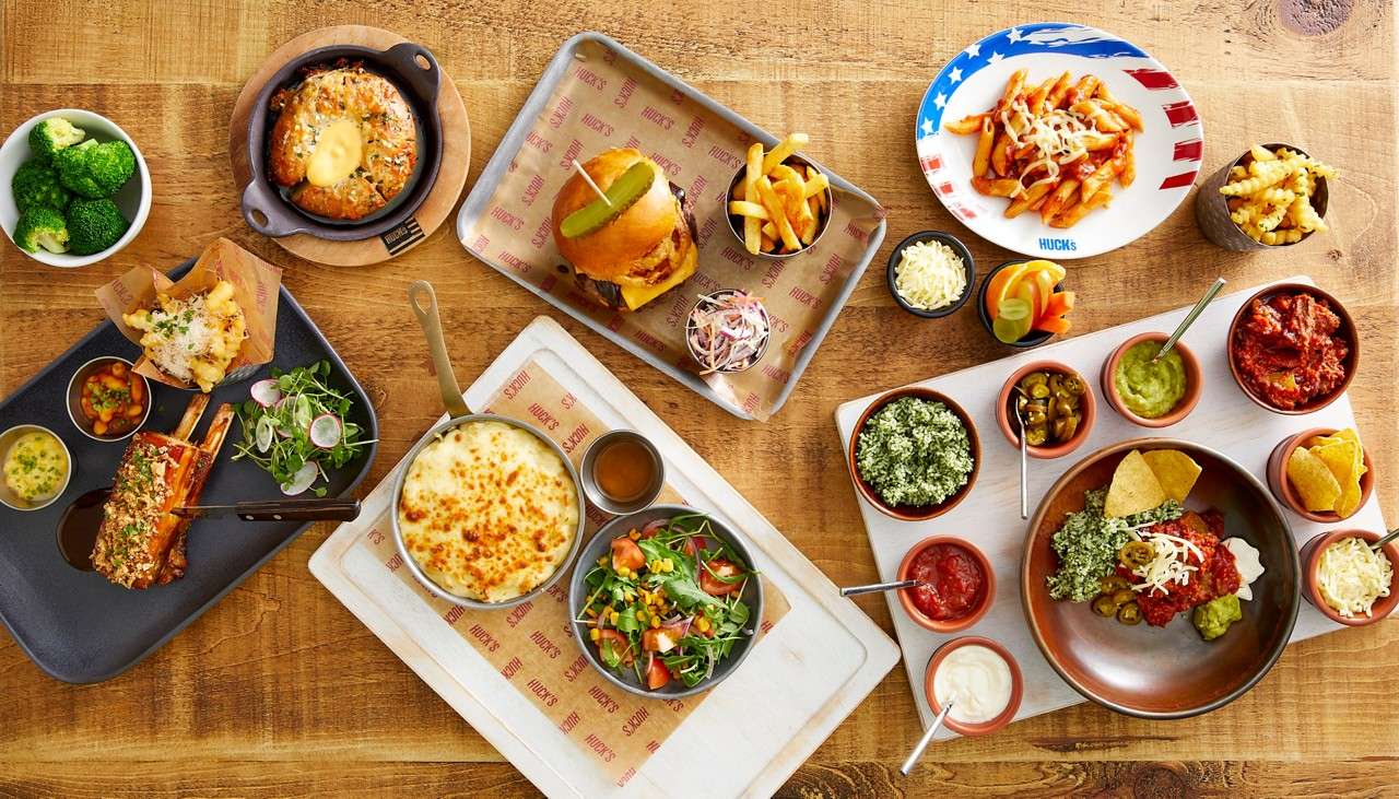 Assorted dishes arranged on a wooden table: cheeseburger with fries, pasta, ribs, nachos with dips, mac and cheese, salads, soft pretzel with sauce, broccoli, and crinkle fries. Text: HUCK’S (repeated), HUCKS.