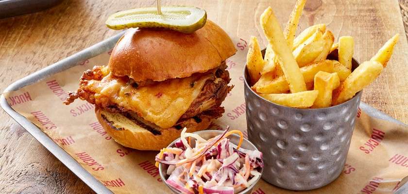 Burger with melted cheese and a pickle spear rests in a bun, accompanied by fries in a metal cup and coleslaw on a tray over paper reading “HUCK’S” repeatedly, on a wooden table.