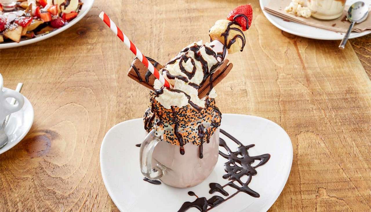 Milkshake overflows with whipped cream and chocolate drizzle, garnished with wafer sticks and a skewer of doughnut and strawberry, sitting on a white plate with chocolate syrup on wooden table.