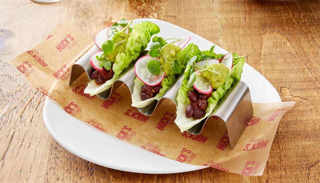 Three lettuce-leaf tacos hold black beans, radish slices, microgreens, and guacamole, resting in metal taco holders on a parchment-lined white plate on a wooden table. Text: HUCK'S Wings (repeated).