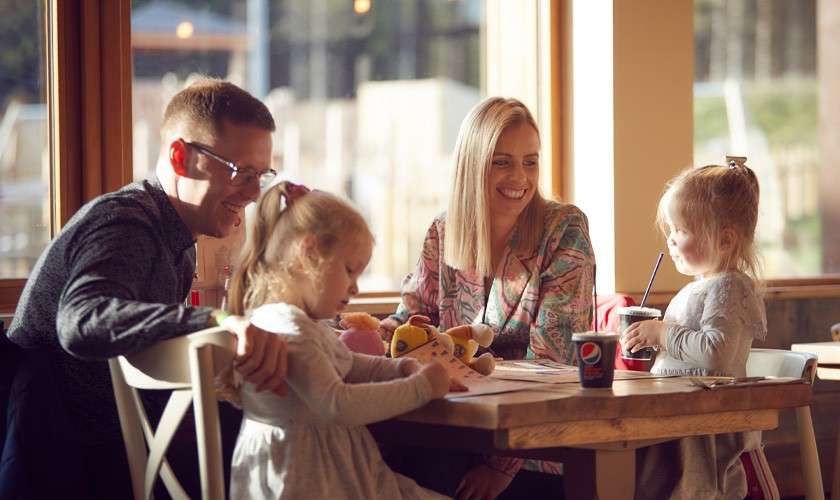 Family sits at wooden table, parents smiling while two young children draw and drink with straws; stuffed toy and cups nearby in a sunlit cafe. Text: pepsi, pepsi.