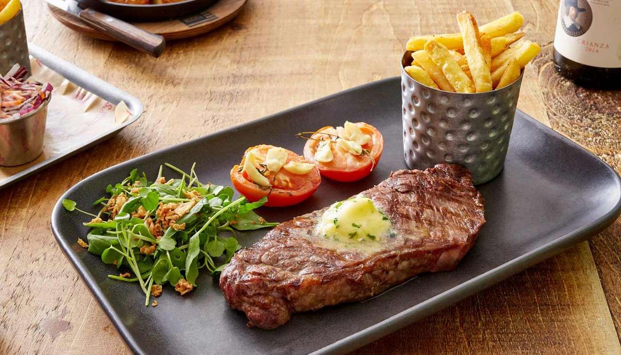 Steak rests topped with melting herb butter, accompanied by cup of fries, grilled tomato halves, and green salad; served on a dark platter on a wooden table with side dishes.
