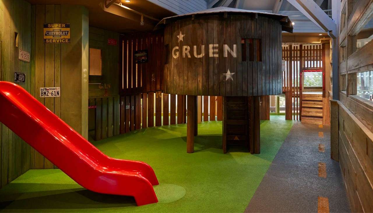 Wooden treehouse stands elevated, accessed by ladder, within an indoor playground; a bright red slide rests nearby on green rubber flooring, surrounded by rustic slatted walls and license-plate decor.Text: GRUEN; SUPER CHEVROLET SERVICE; TEXAS BMZ-61N; EXIT.