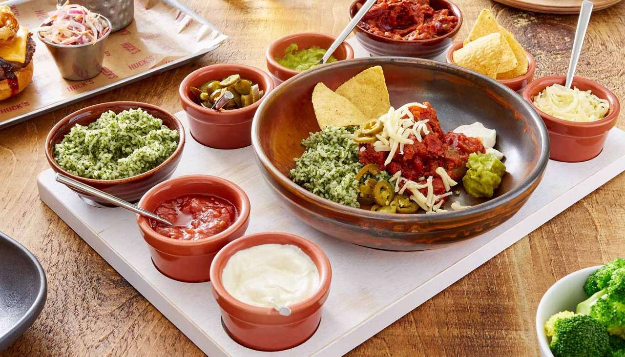 Bowl of nachos holds salsa, shredded cheese, guacamole, jalapeños, and chips, surrounded by ramekins of green herby mixture, salsa, sour cream, olives, and more chips on a wooden table; tray paper reads: “HUCK’S”.