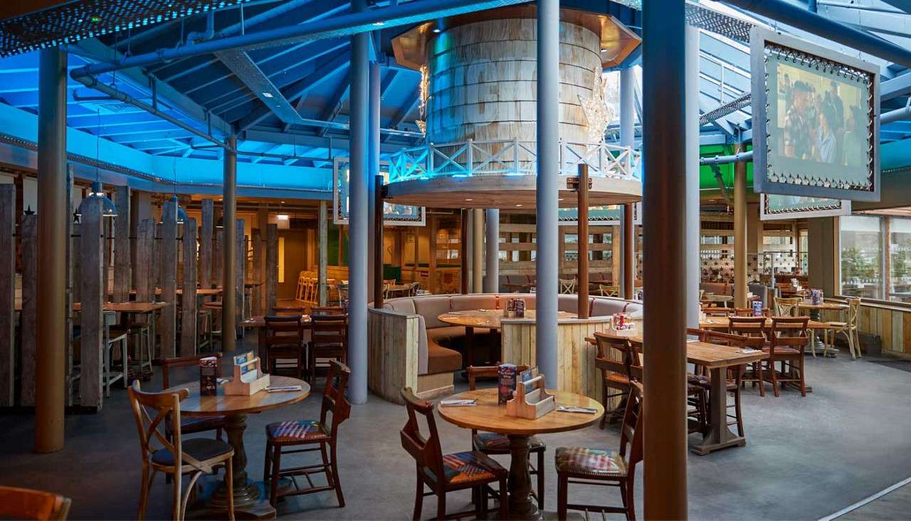 Dining tables and a circular booth seat patrons, arranged neatly beneath a blue-lit, exposed-beam roof, with columns, hanging screens, and rustic wood accents in a spacious restaurant interior.