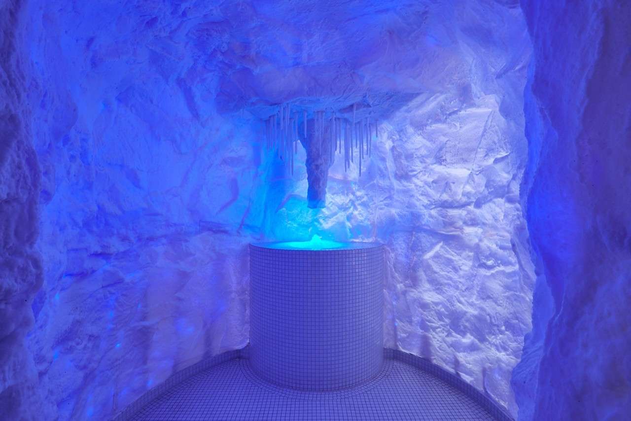Cylindrical tiled basin glows blue while water or light pours from hanging icicles above, set within a cave-like, ice-themed room with textured white walls and curved tiled floor.