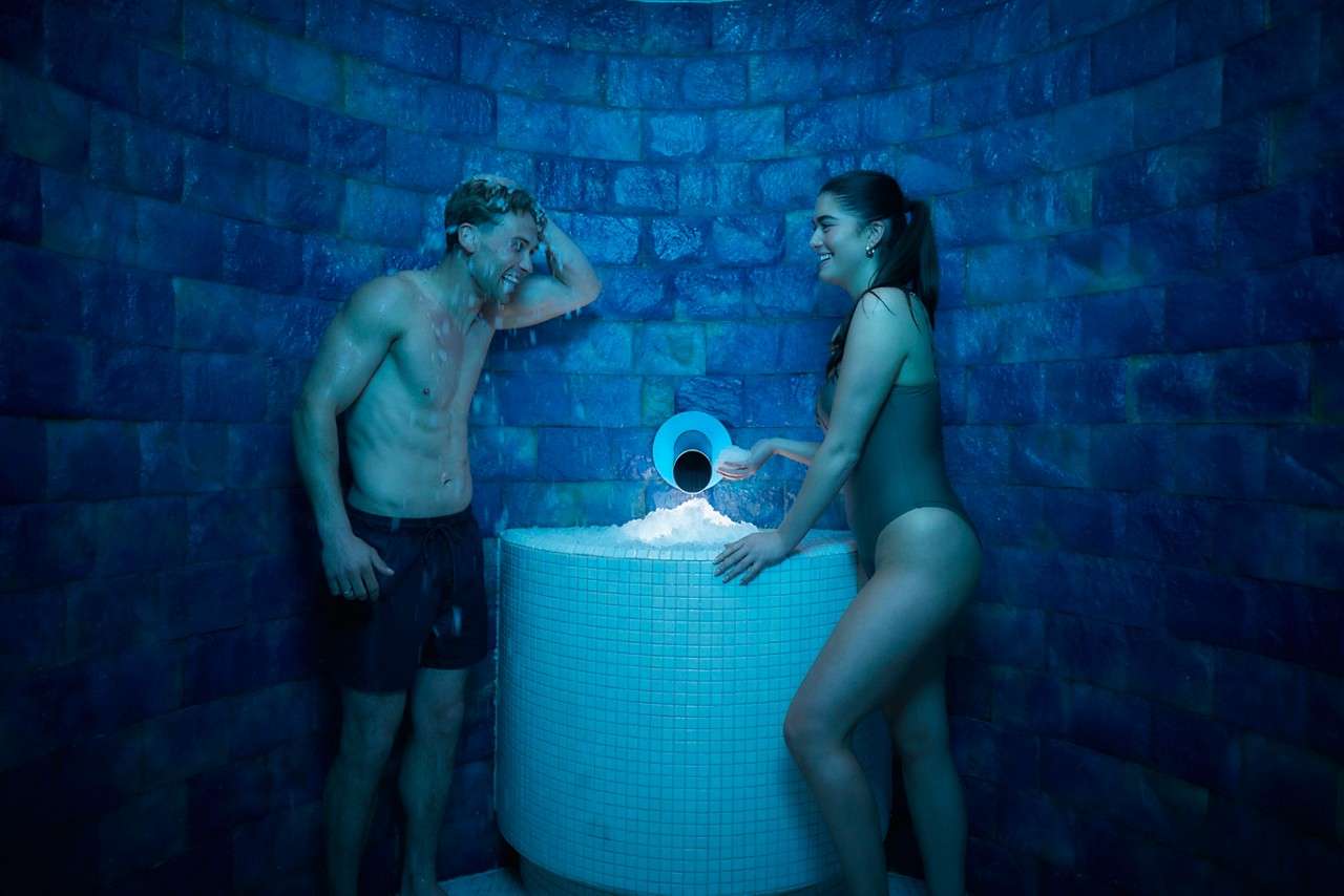 Two swimsuit-clad adults scoop crushed ice from a tiled pedestal, smiling and rubbing it on themselves, inside a blue-lit, round stone chamber.