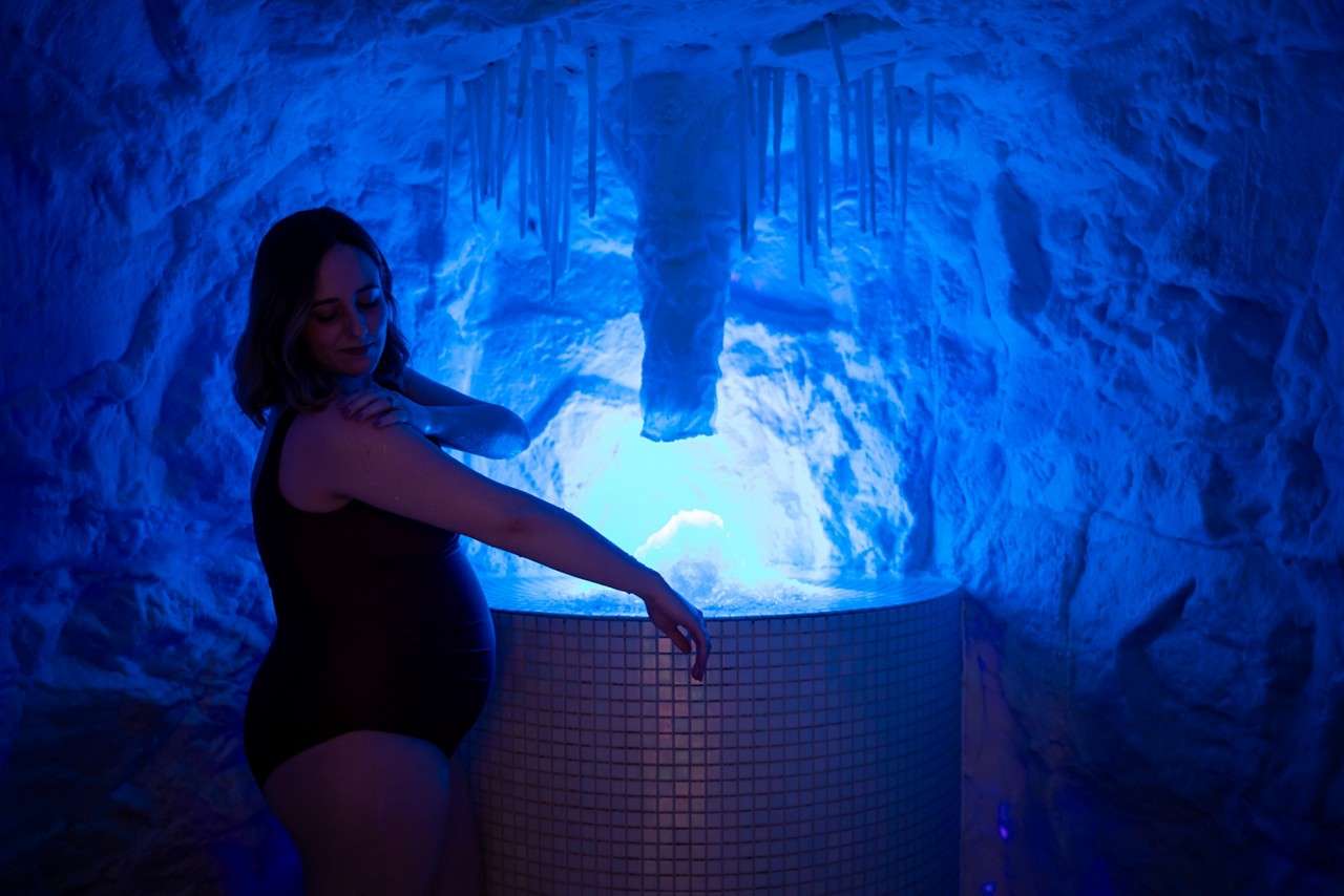 A pregnant person in a dark swimsuit rests one arm on a tiled round fountain, gently touching their shoulder, in a blue-lit cave-like room with icicle shapes overhead.