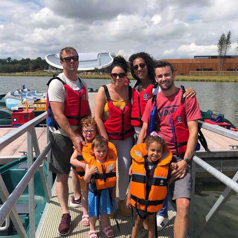 Group of adults and children wear life jackets, standing and smiling on a dock, preparing for paddle boats on a lake; shirt text: VANS; child’s life vest label: EVIE.