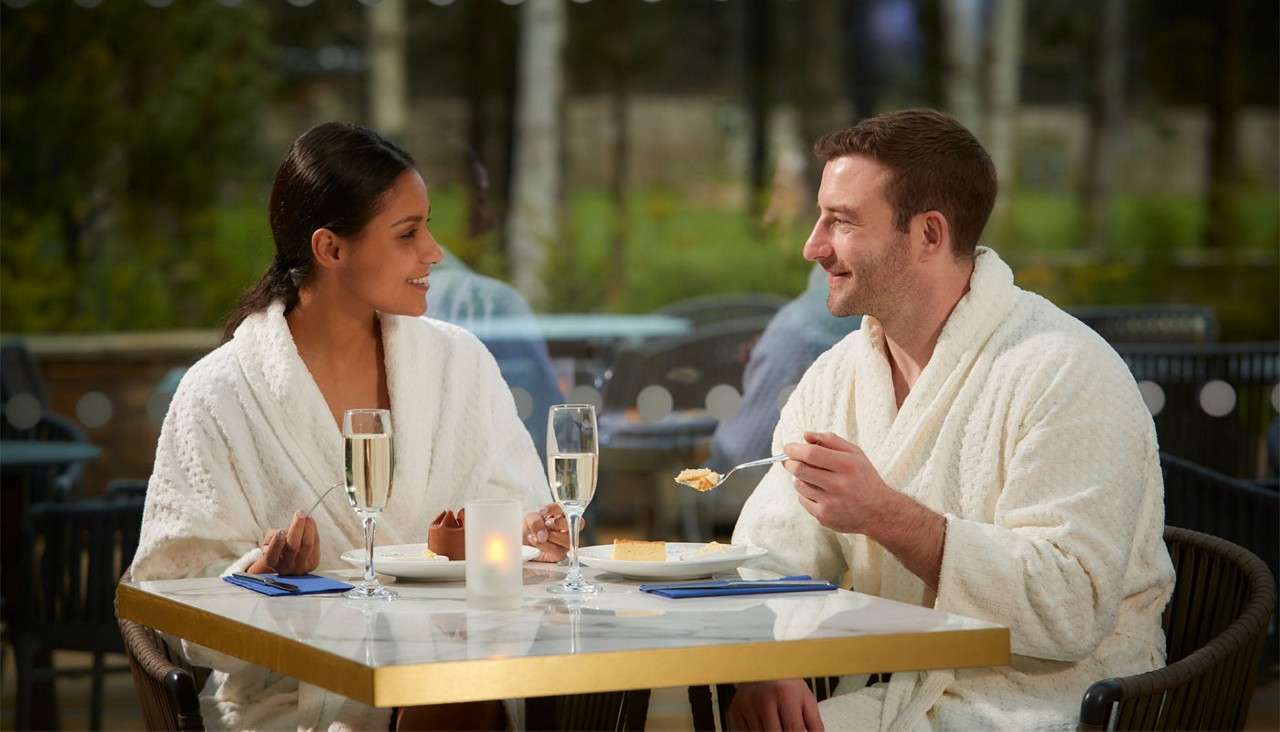 Two people in white bathrobes eat dessert and converse, holding utensils beside flutes of sparkling drink on a candlelit table, seated on an outdoor patio with greenery and blurred background.
