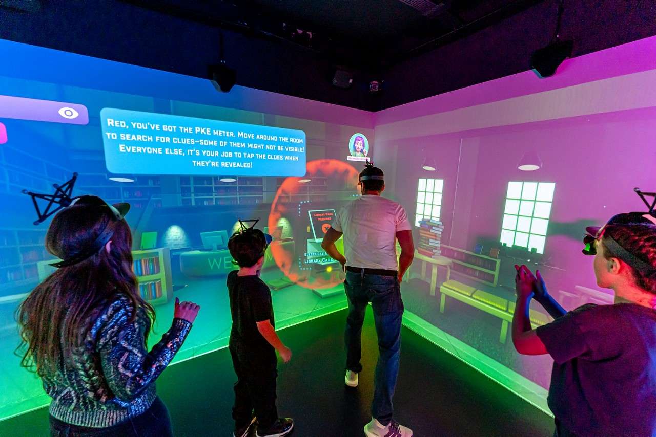 Players wearing headsets gesture and move, interacting with a wall-sized digital game in a room. On-screen text: "RED, YOU'VE GOT THE PKE METER. MOVE AROUND THE ROOM TO SEARCH FOR CLUES—SOME OF THEM MIGHT NOT BE VISIBLE! EVERYONE ELSE, IT'S YOUR JOB TO TAP THE CLUES WHEN THEY'RE REVEALED!" Visible sign: "WELCOME".