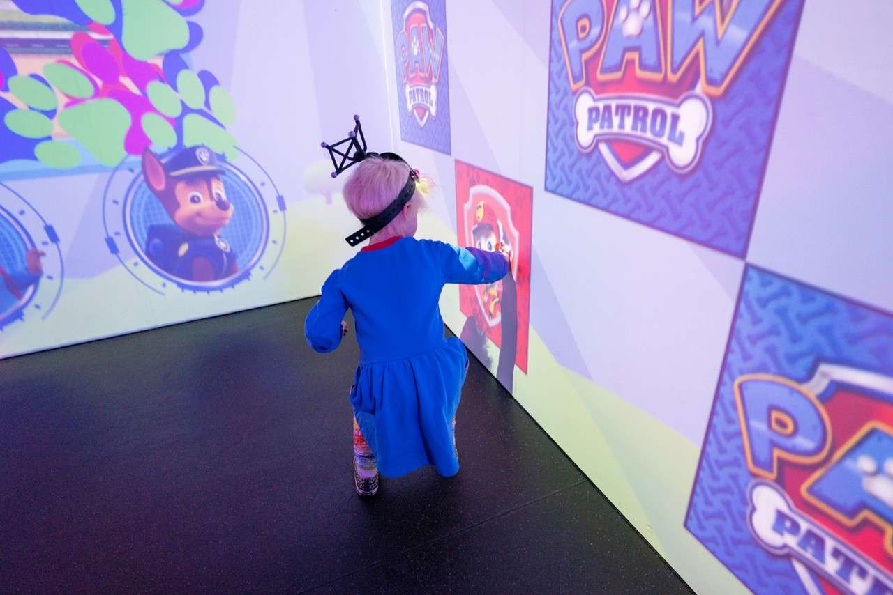 Child in a blue dress wearing a head-tracking rig touches a wall projection, interacting with cartoon panels in a play space. Text: “PAW Patrol”, “PAW Patrol”, “PAW”.