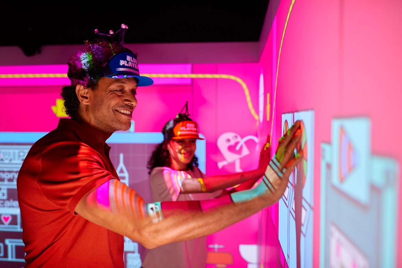 Two participants press their hands on a neon interactive wall, smiling as projected graphics respond; they wear visors labeled: BLUE PLAYER and RED PLAYER, inside a colorful arcade-like environment.