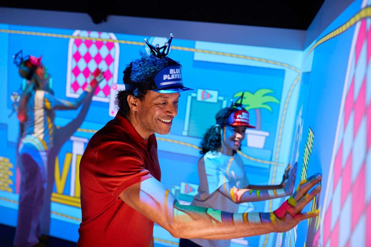Players wearing tracking caps press hands against an interactive, cartoon-projected wall, engaging in a collaborative game inside a dark, neon-lit immersive room. Text: BLUE PLAYER; RED PLAYER; IMMERSIVE GAMEBOX.