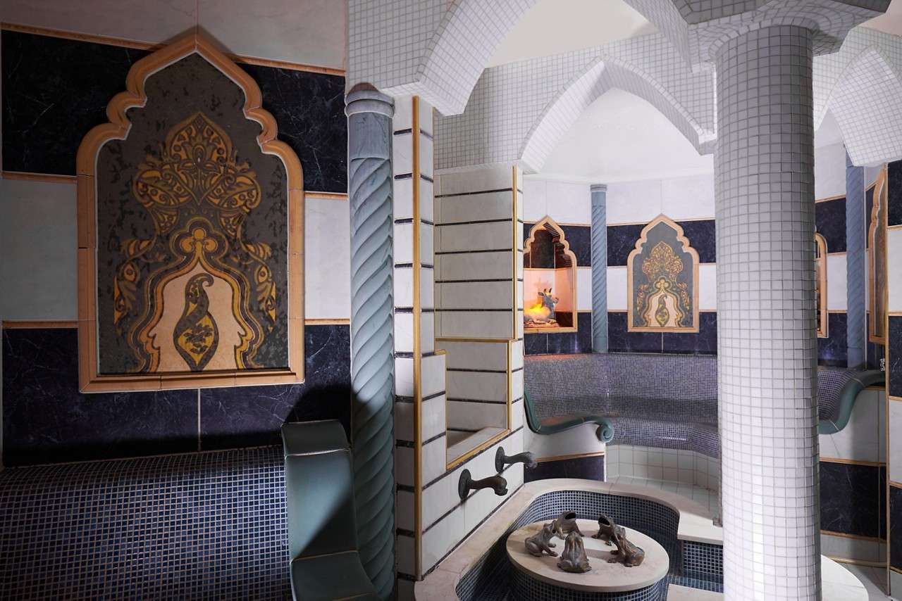 Ornate tiled hammam room—curved bench seating lines the walls, water taps feed a small central basin with figurines—surrounded by arched niches, patterned panels, spiral columns, and vaulted, mosaic-covered ceilings.