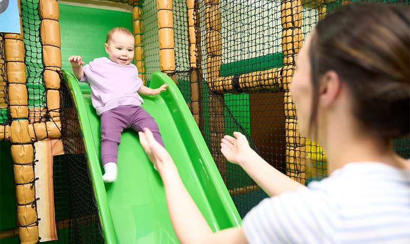 Baby slides down a bright green slide, smiling, while an adult with outstretched arms waits to catch them in an indoor soft-play area enclosed by netting and padded structures.