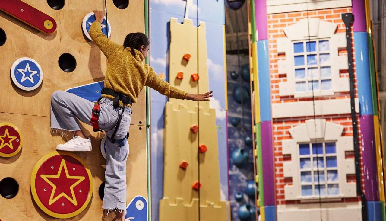 Climber reaches sideways while ascending a colorful indoor climbing wall, secured by a harness; themed walls display stars, orange holds on a castle-like panel, and a brick building facade.