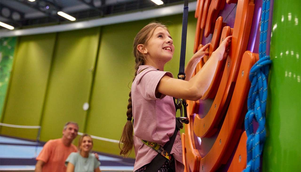 Girl in harness climbs a colorful indoor climbing wall, gripping orange holds. In the background, two adults watch from below in a spacious, green-walled recreation center with overhead lights.