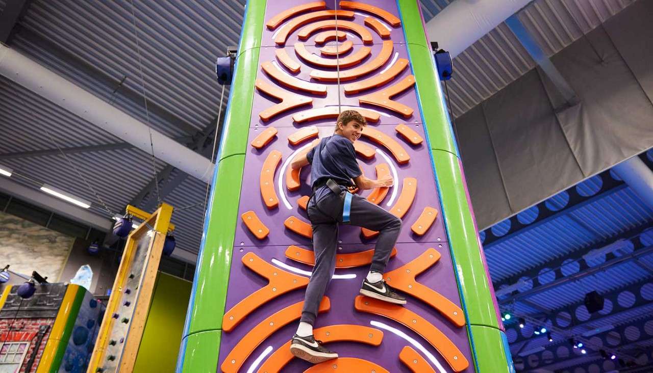 A harnessed teenager climbs a colorful indoor climbing wall with orange curved holds, looking back toward the camera, inside a large gym with metal ceiling, lights, and other climbing structures.