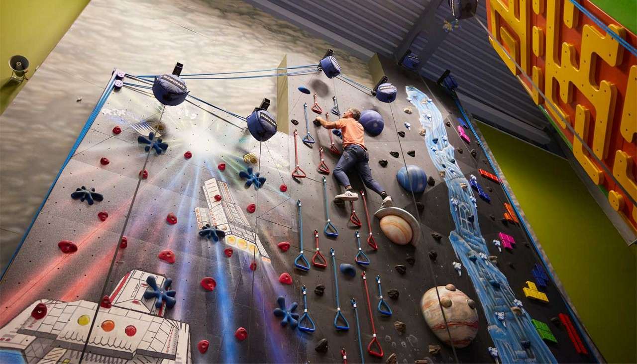 Climber scales a colorful indoor rock wall, gripping holds and rope loops, while planet-shaped volumes and a space-themed mural surround them inside a bright, high-ceilinged climbing gym.